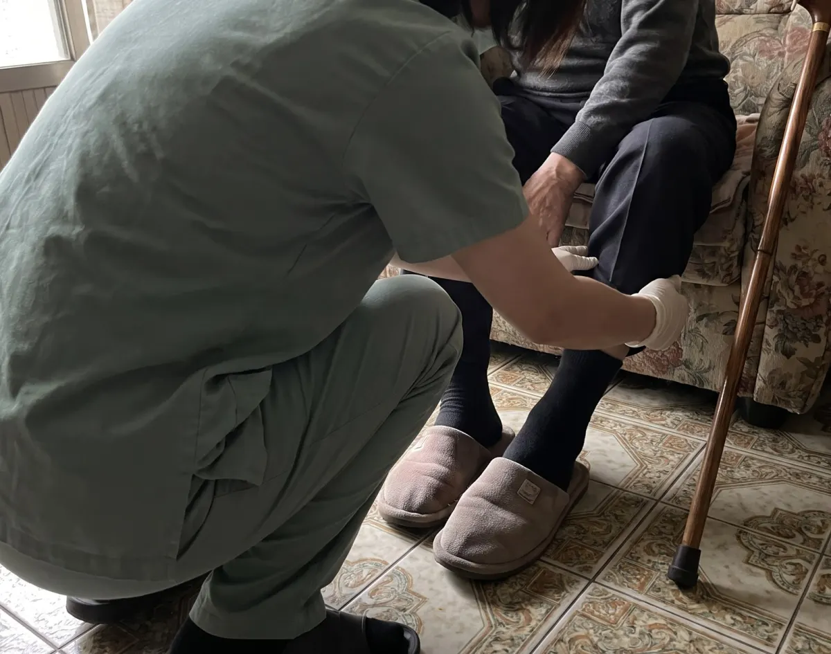 阿爾瑪護理師蹲下幫長輩穿拖鞋 / An Alma nurse kneels to help an older woman with her slippers